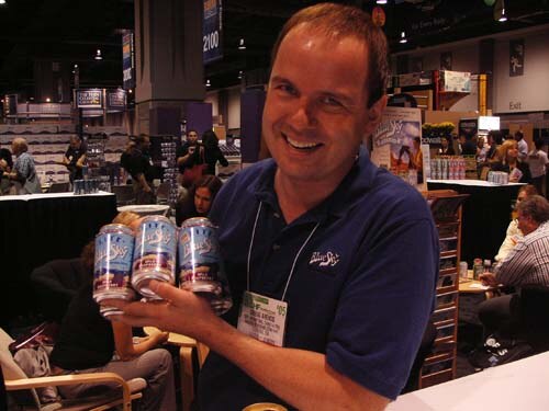 A smiling man in a navy Blue Sky polo with a badge holds up three cans of Blue Sky soda toward the camera, with other booths and attendees behind him.
