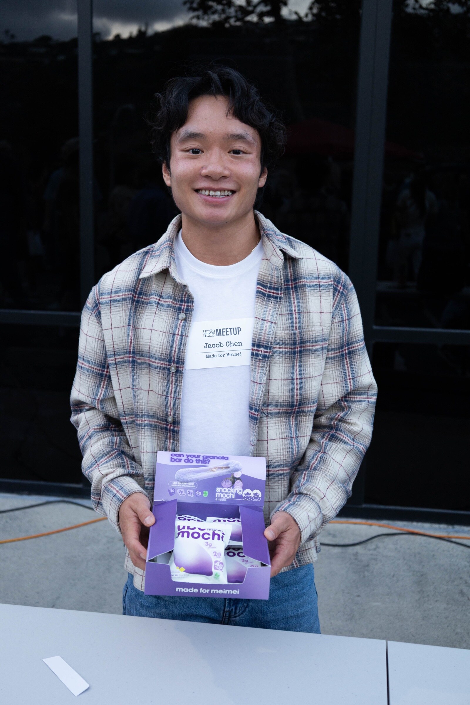 Smiling young man in a plaid shirt holds an open box of Made for Meimei ube snacking mochi.
