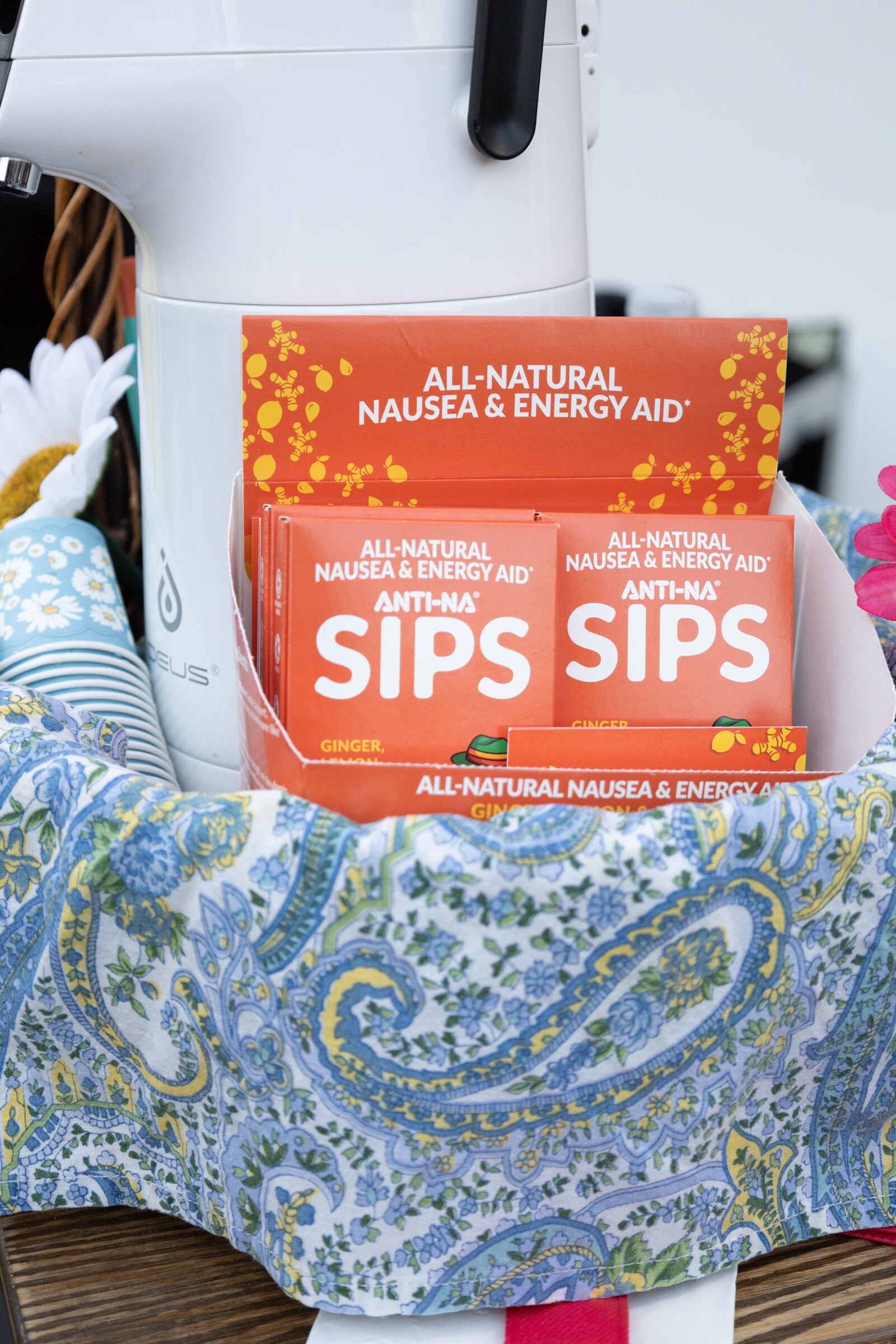 Orange boxes of Anti-Na Sips all-natural nausea and energy aid in a basket lined with blue paisley fabric.