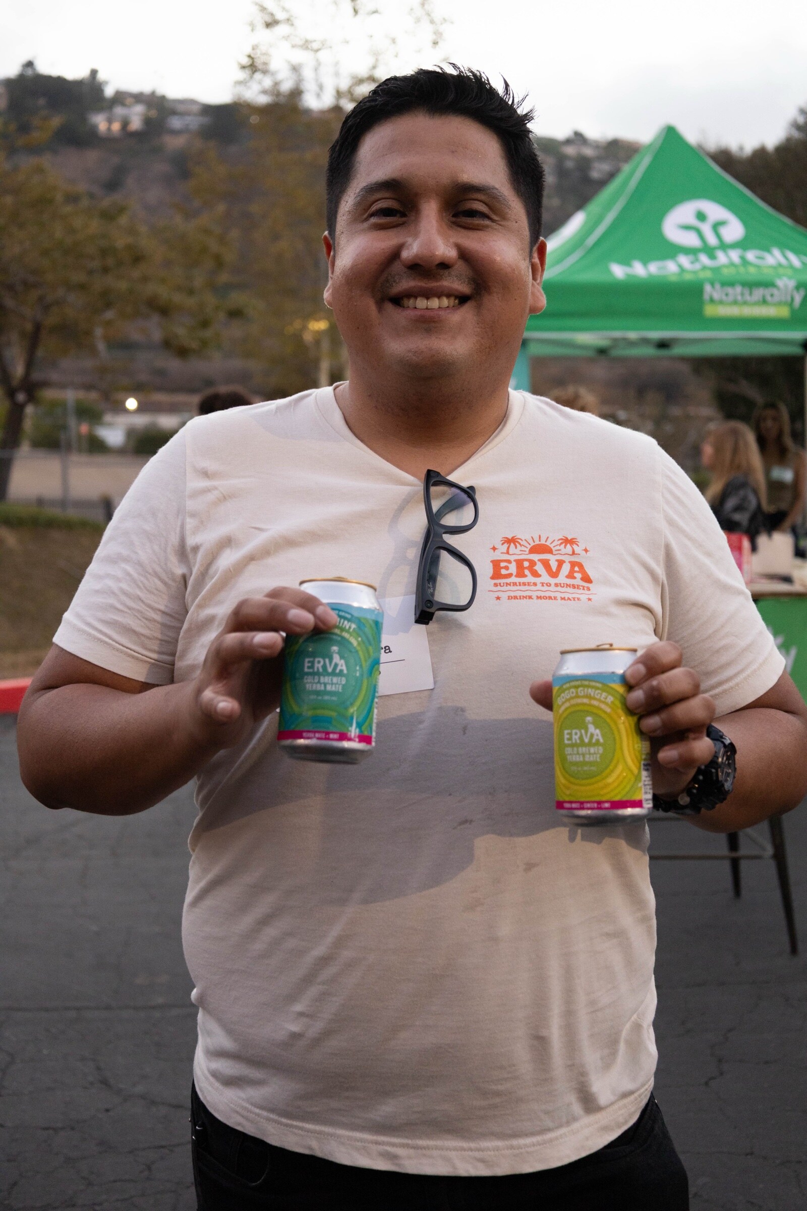 Smiling man in an Erva T-shirt holds up two cans of Erva cold-brewed yerba mate, with the Naturally San Diego tent behind.