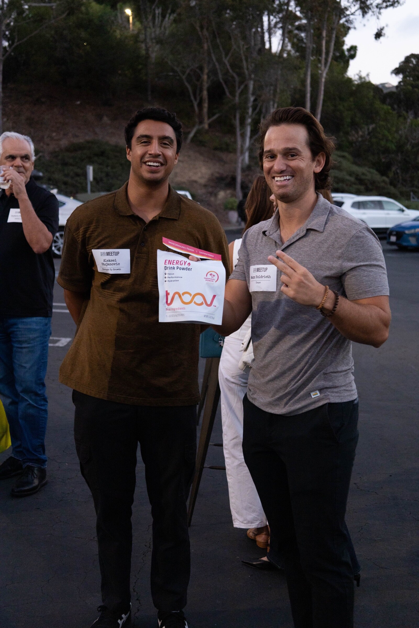 Two smiling men pose in a parking lot, one holding a pouch of Troov Tropical Punch energy drink powder.