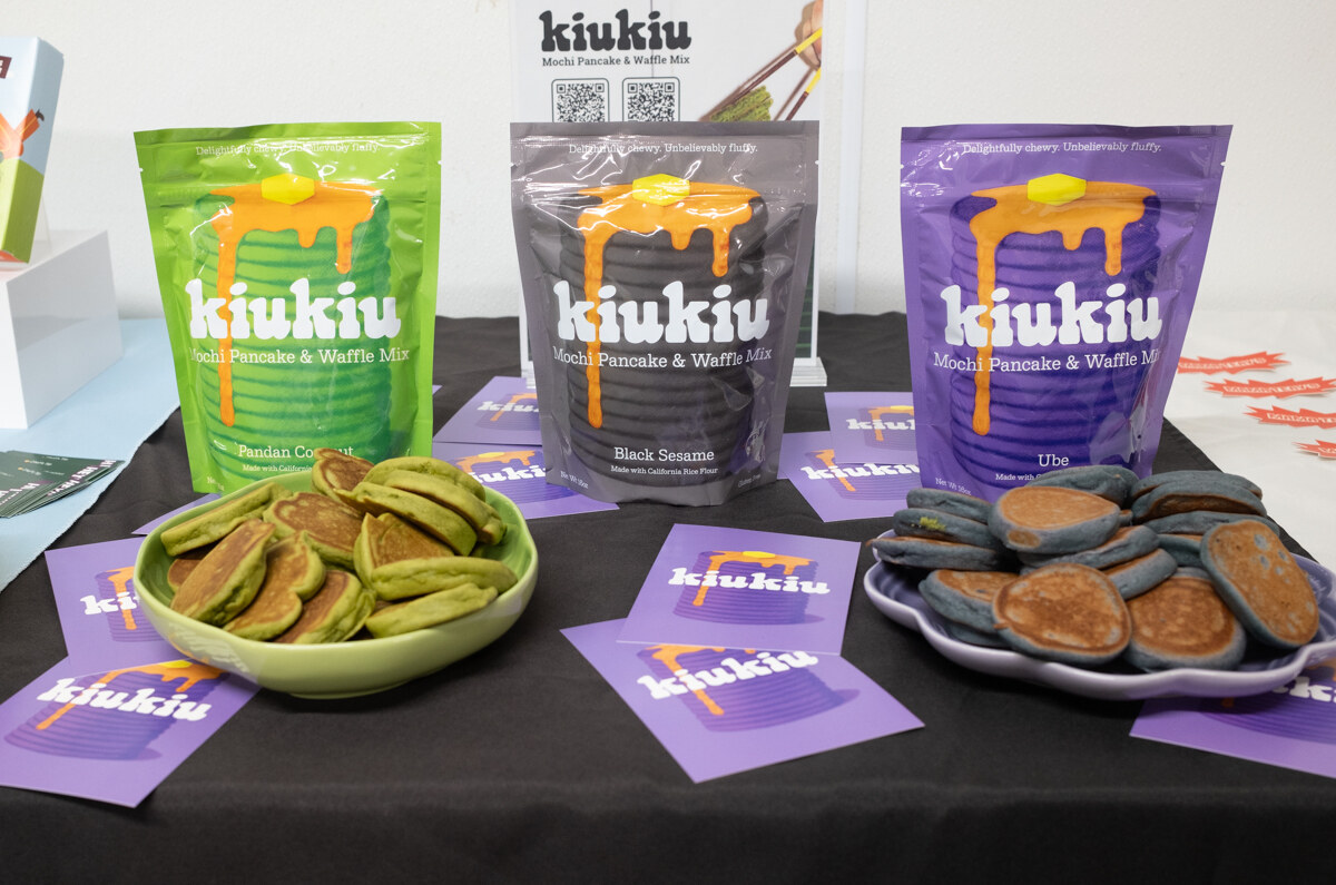 Bags of kiukiu pancake mix in green, black and purple beside plates of colored pancakes.