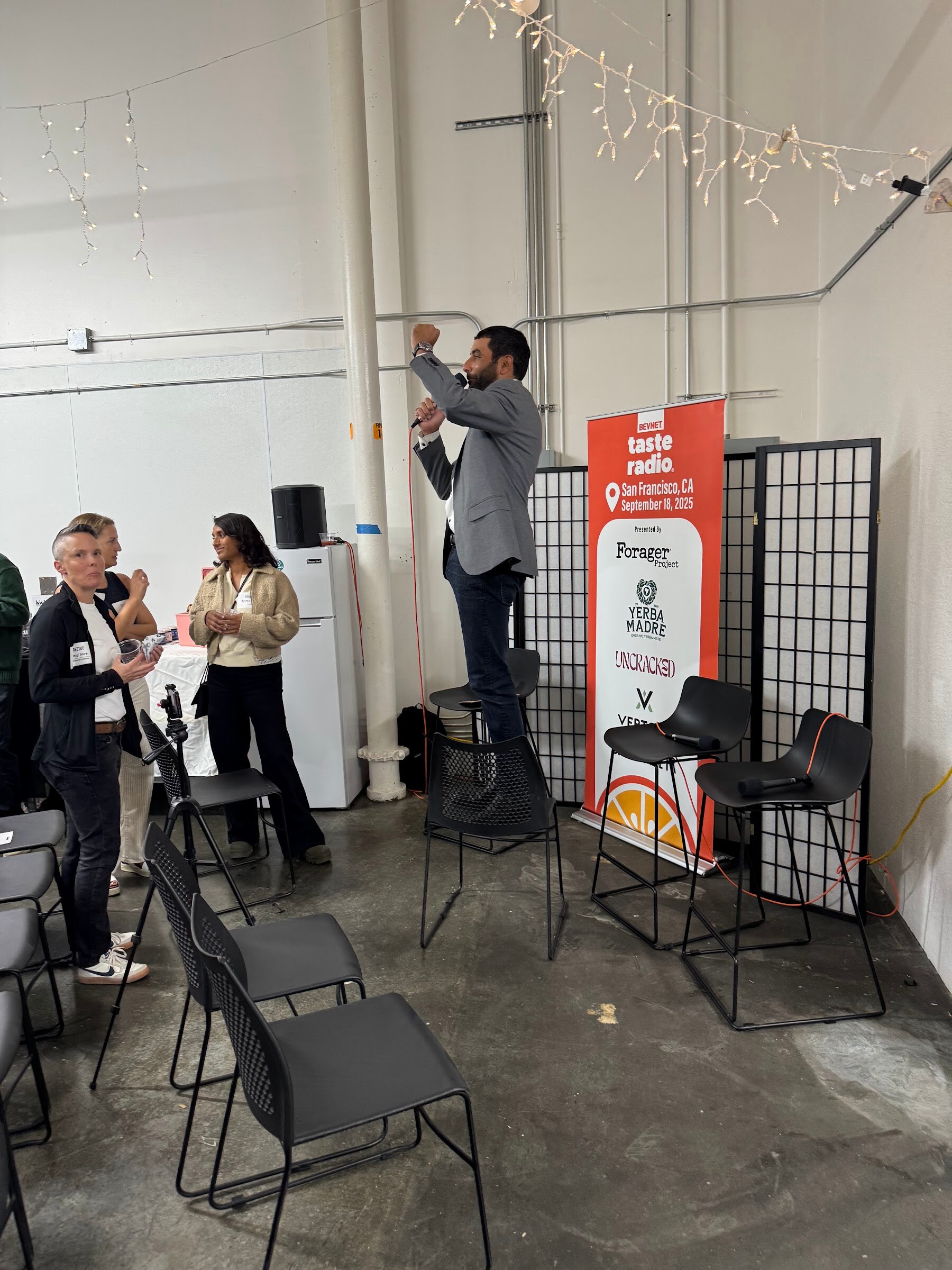 Man in a gray blazer standing by a banner and empty chairs, with a few guests nearby.