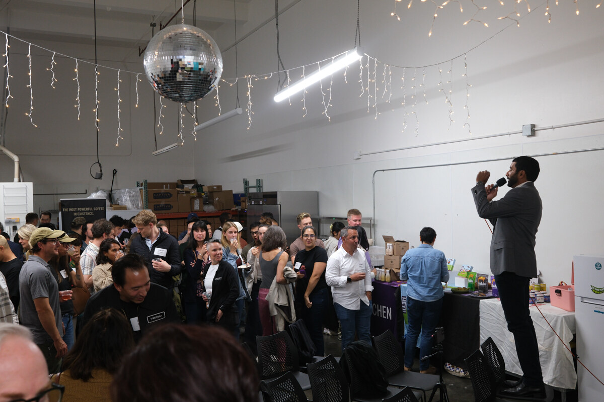 Man in a gray blazer holding a microphone in a crowded warehouse room hung with string lights and a disco ball.