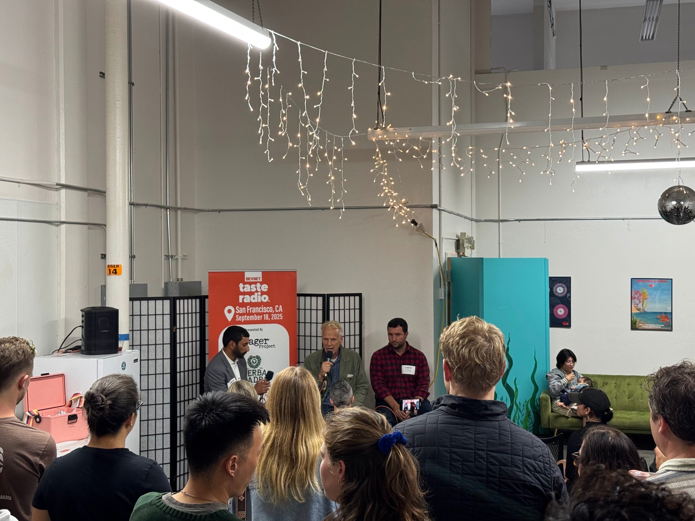 Three people on stools in front of the banner, seen past rows of attendees.
