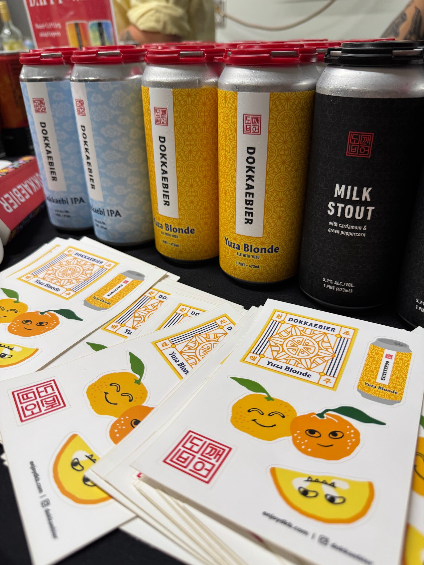 Cans of Dokkaebier beer lined up above stacks of stickers on a black tablecloth.