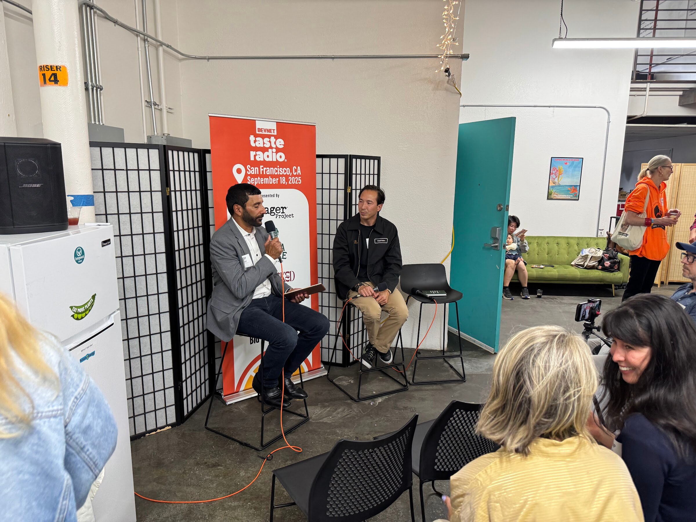 Two men seated on stools in front of the Taste Radio San Francisco banner with an audience nearby.