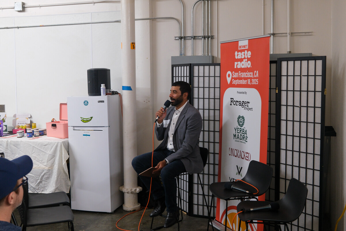 Man in a gray blazer holding a microphone, seated beside the banner and a white mini fridge.