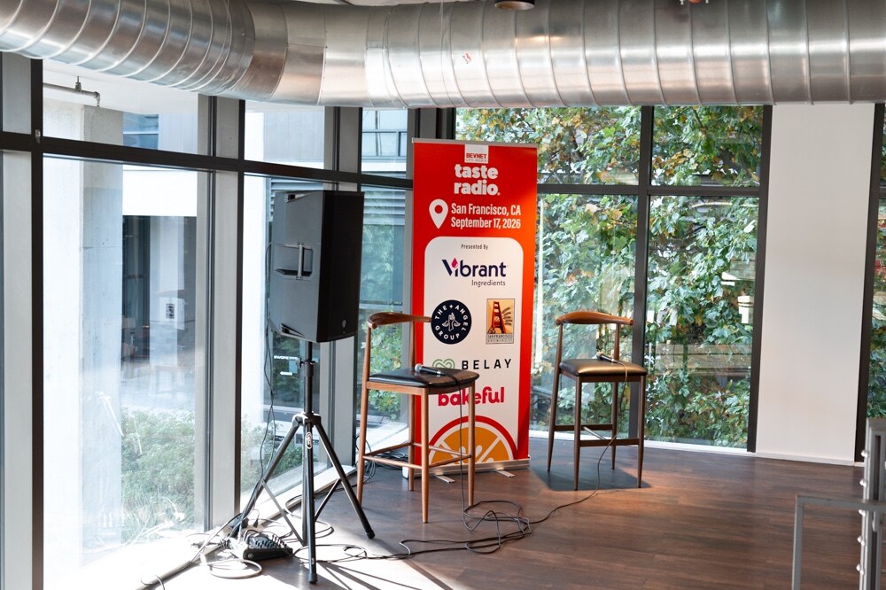 Two stools and a speaker beside the Taste Radio San Francisco September 17, 2026 banner in front of tall windows.