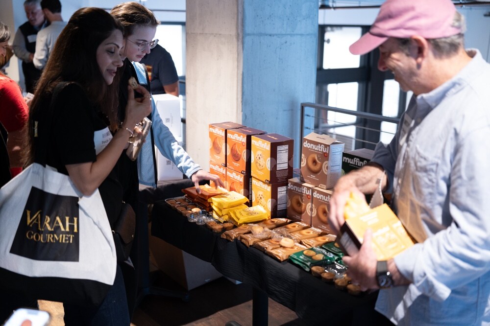 Attendees, one carrying a Varah Gourmet tote, browse a table stacked with Donutful and Muffinful boxes.