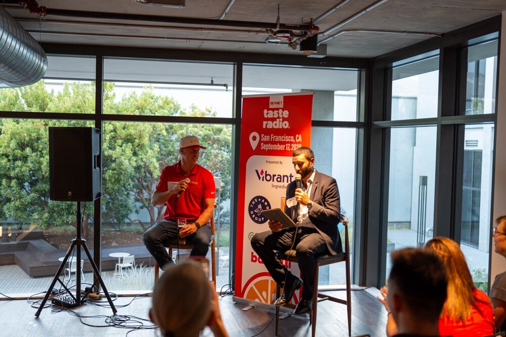 Man in a red shirt and cap and the host in a suit sit on stools beside the Taste Radio San Francisco banner.