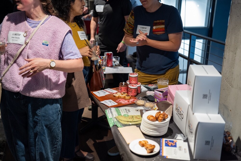 Attendees, one in a pink vest, sample from a table with boxes and plates.