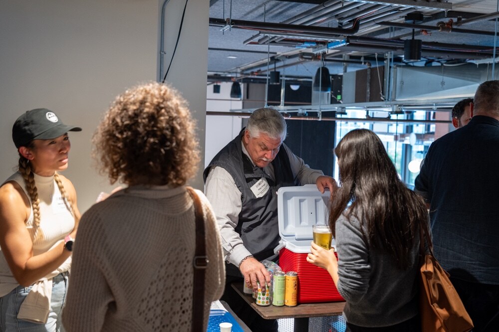 Man in a vest pulls cans from a red cooler as attendees watch.