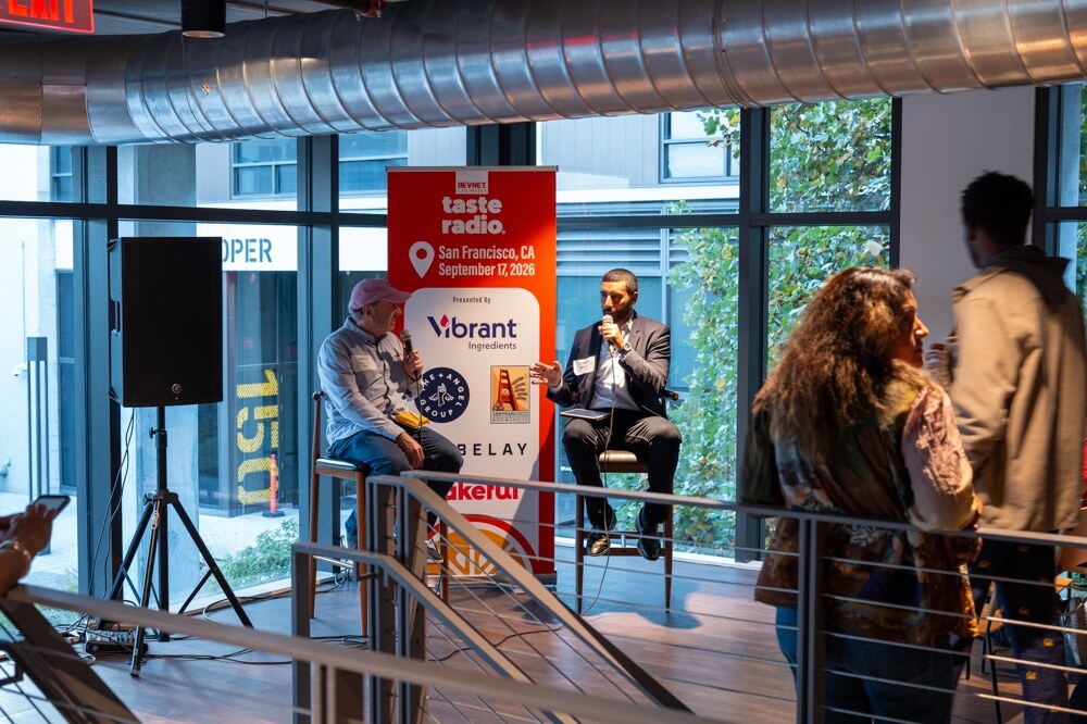 Host and guest on stools by the banner, with attendees watching from behind a railing.