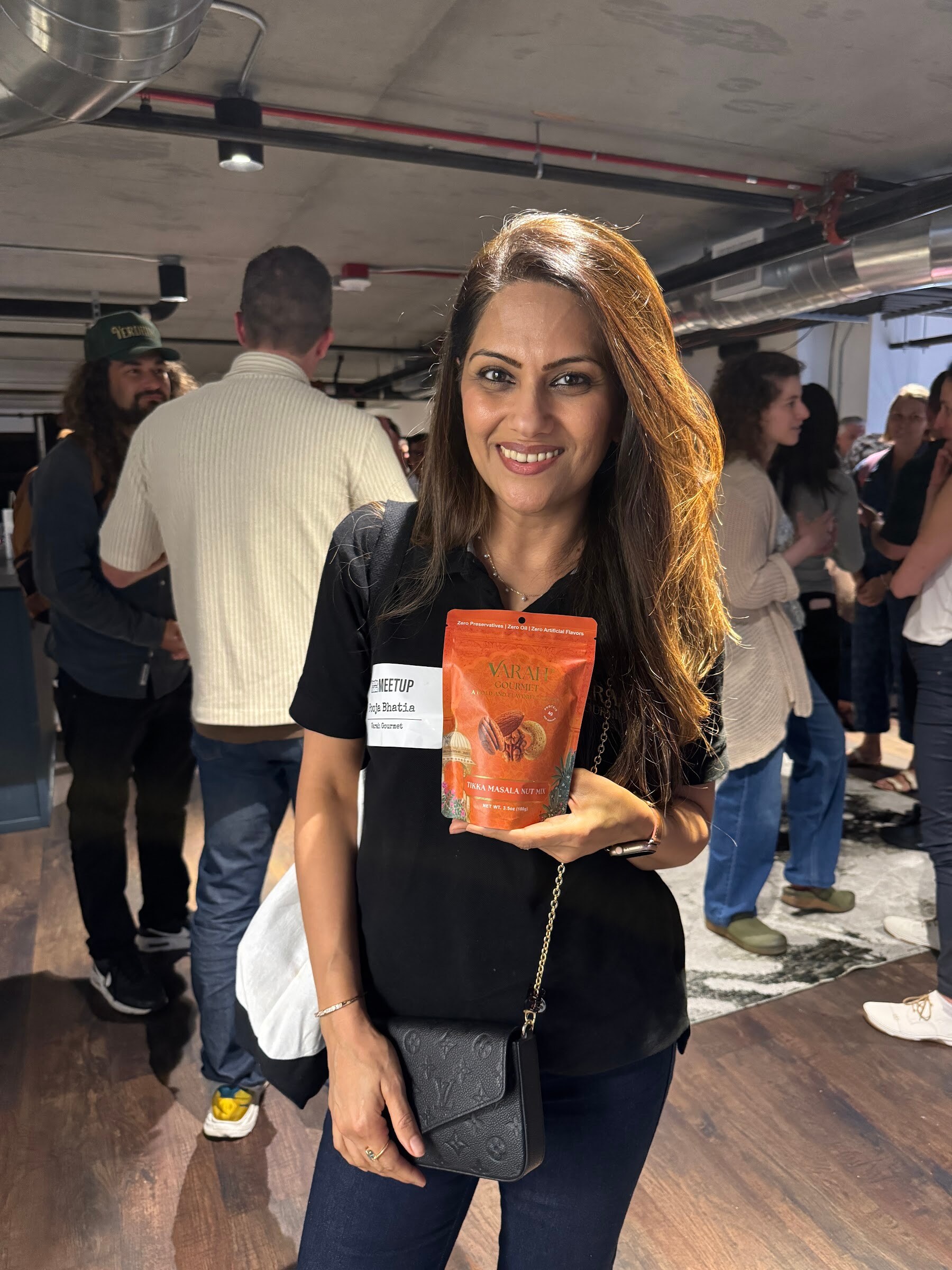 Smiling woman holds a pouch of Varah Gourmet Tikka Masala nut mix in a crowded room.