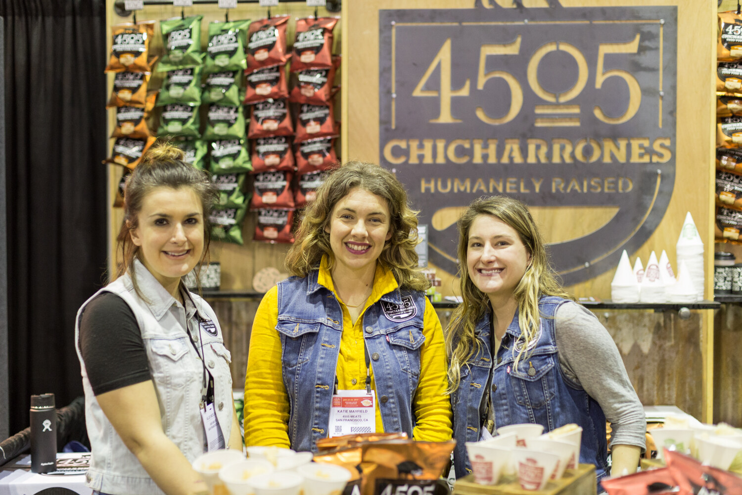 Three smiling women stand at a counter with sample cups in front of a wooden wall with a 4505 Chicharrones Humanely Raised sign and hanging bags of snacks.