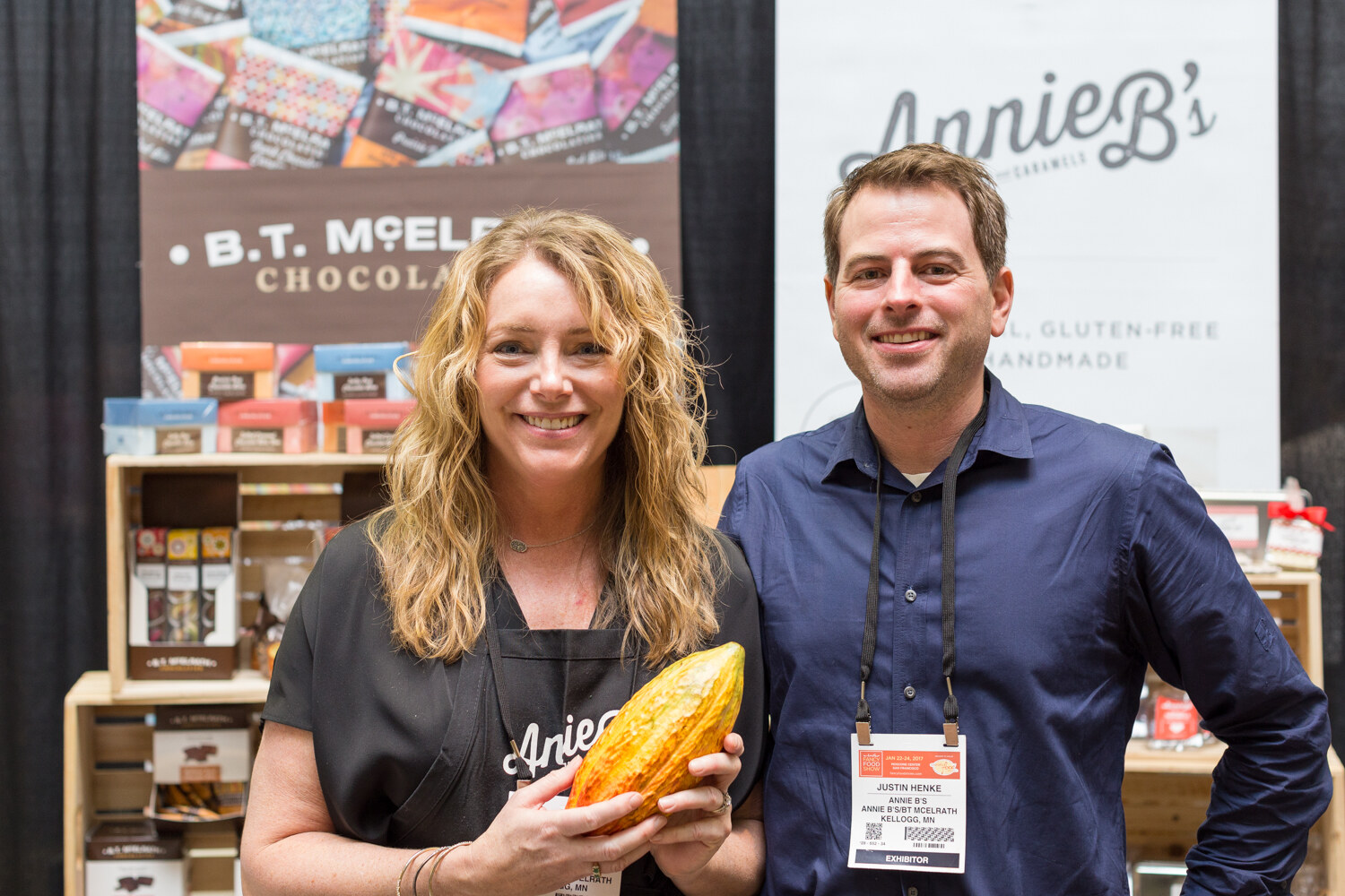 A woman holding a yellow cacao pod and a man in a blue shirt stand in front of B.T. McElrath Chocolate displays and an Annie B's banner.