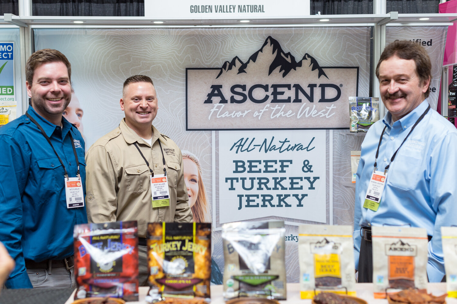 Three men with exhibitor badges stand behind bags of jerky in front of an Ascend Flavor of the West All-Natural Beef and Turkey Jerky sign.
