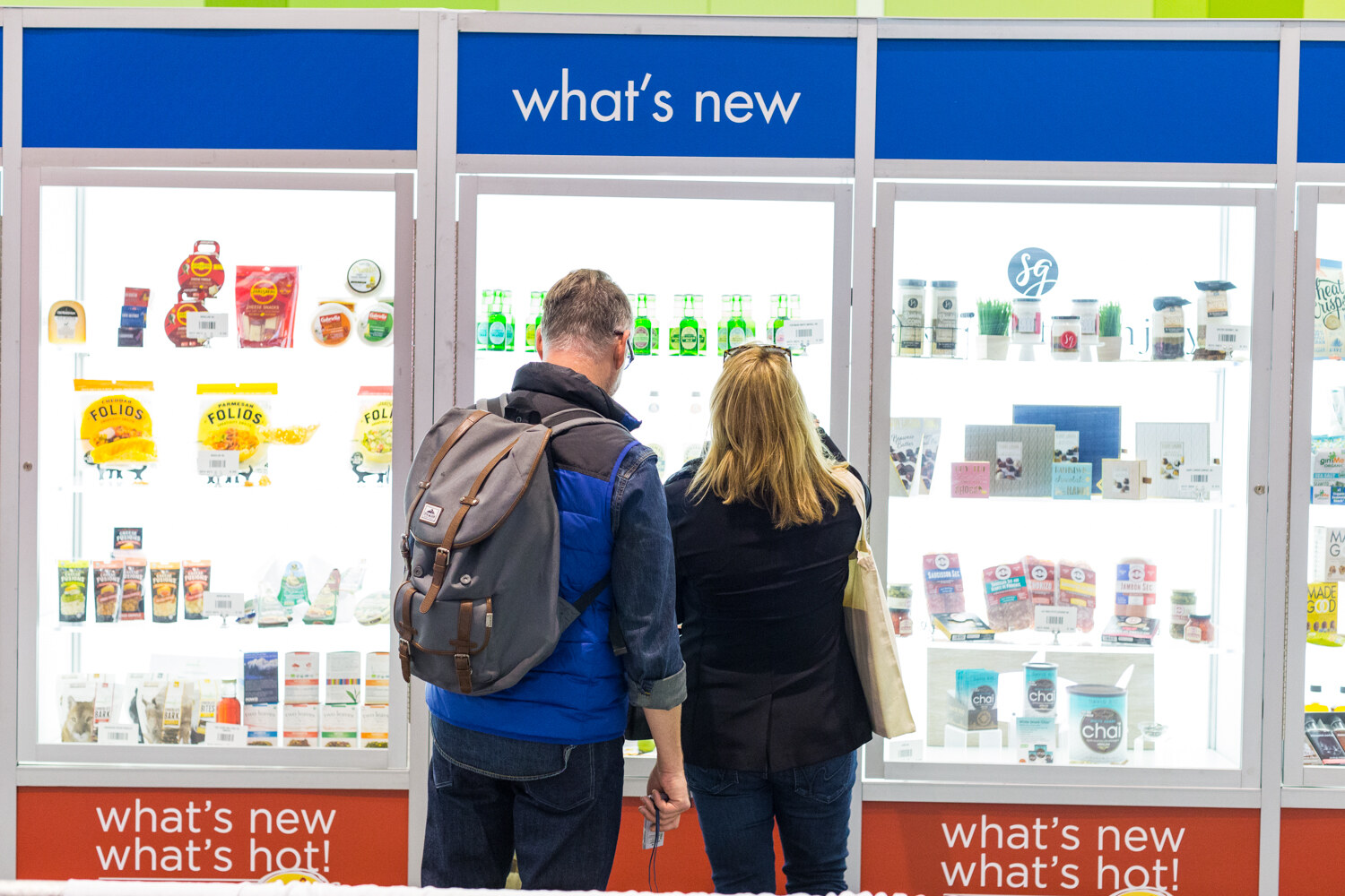 Two people seen from behind look into illuminated glass display cases of packaged foods under blue signs reading what's new.