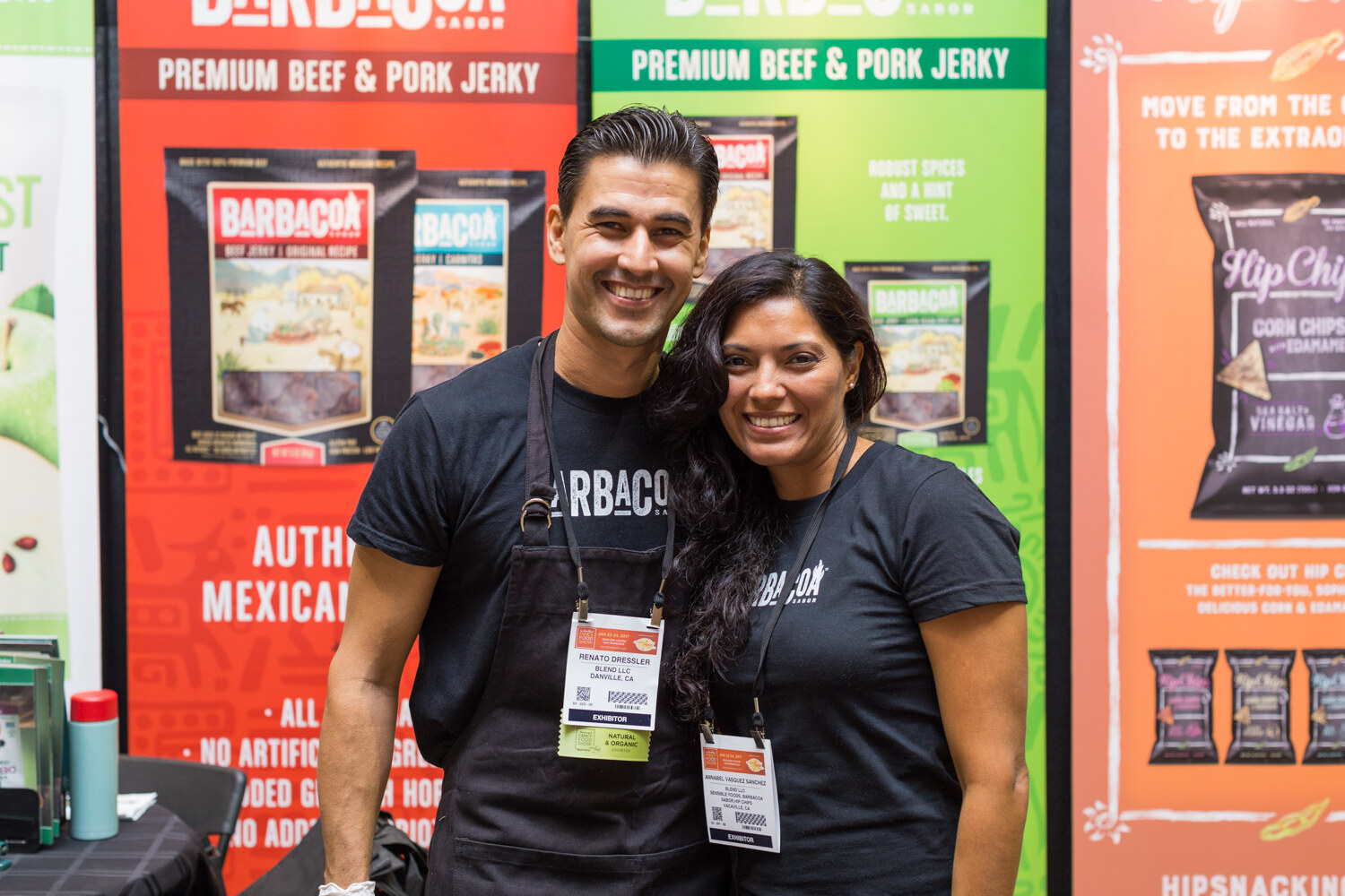 A man and a woman in black Barbacoa shirts and aprons stand in front of red and green banners reading Premium Beef and Pork Jerky.