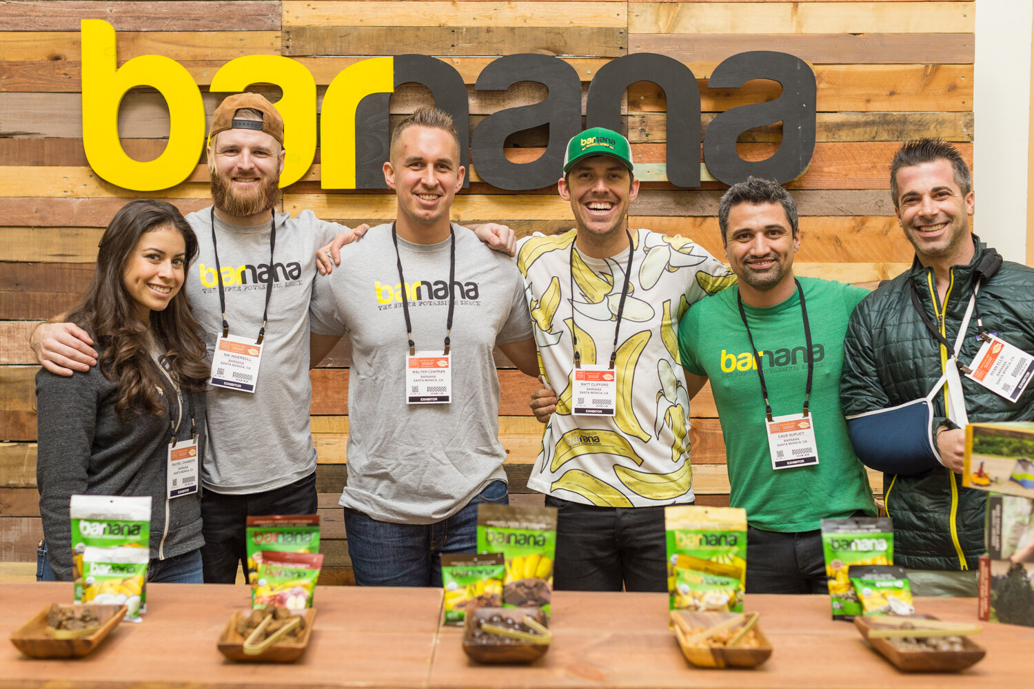 Six people stand shoulder to shoulder at a wooden counter set with banana snack bags, under a large yellow and gray barnana sign.