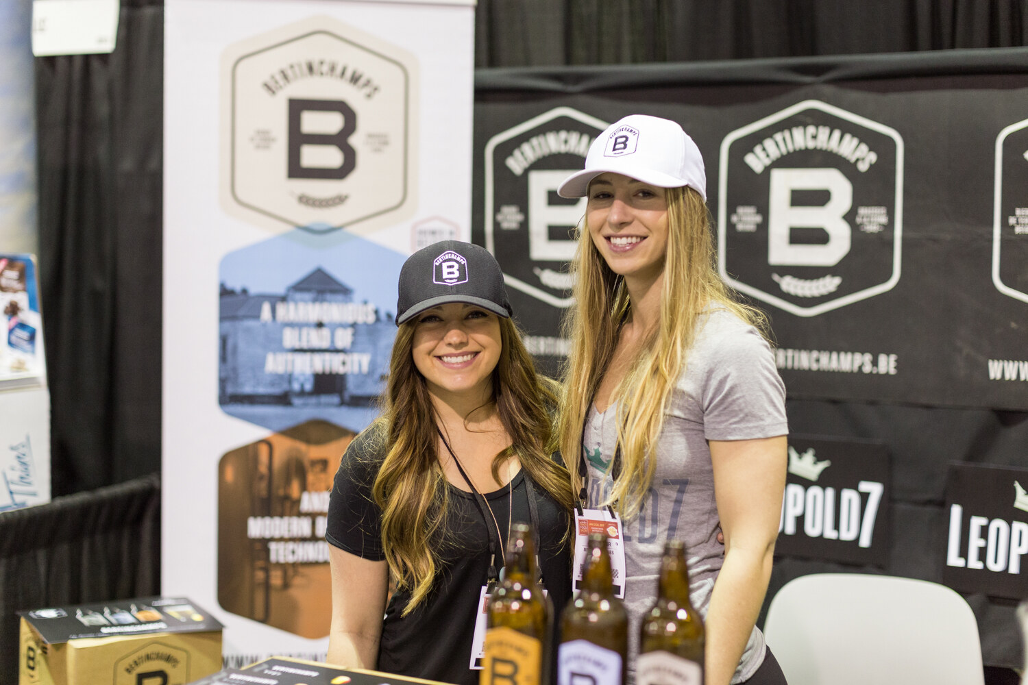 Two smiling women in baseball caps stand behind amber bottles in front of Bertinchamps and Leopold7 banners.