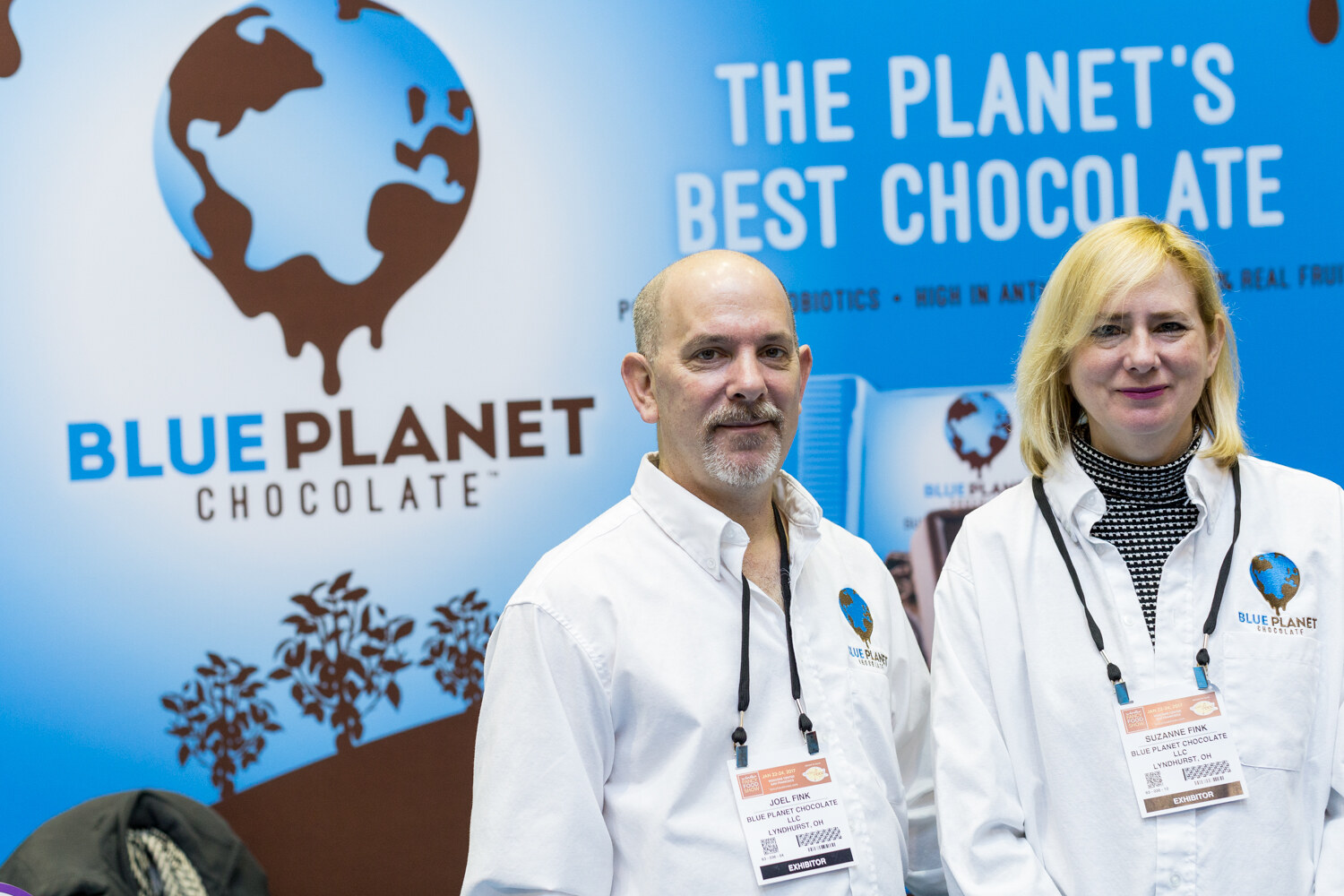 A man and a woman in white jackets with a Blue Planet Chocolate logo stand in front of a blue banner reading The Planet's Best Chocolate.
