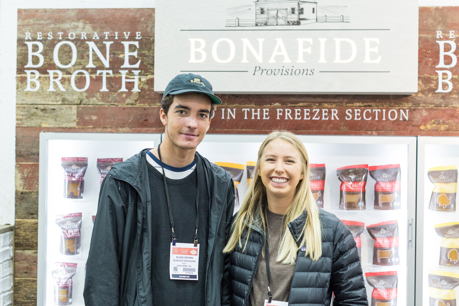 A young man and a young woman stand in front of a rustic wood wall reading Restorative Bone Broth and a freezer case of Bonafide Provisions packages.
