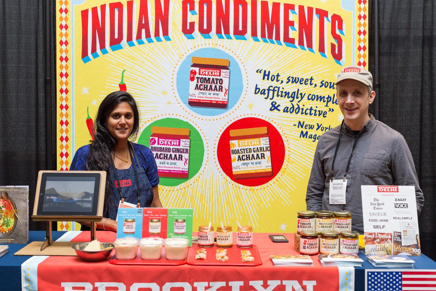 A woman and a man stand behind a table of Brooklyn Delhi Tomato Achaar and Roasted Garlic Achaar jars in front of a yellow Indian Condiments backdrop.