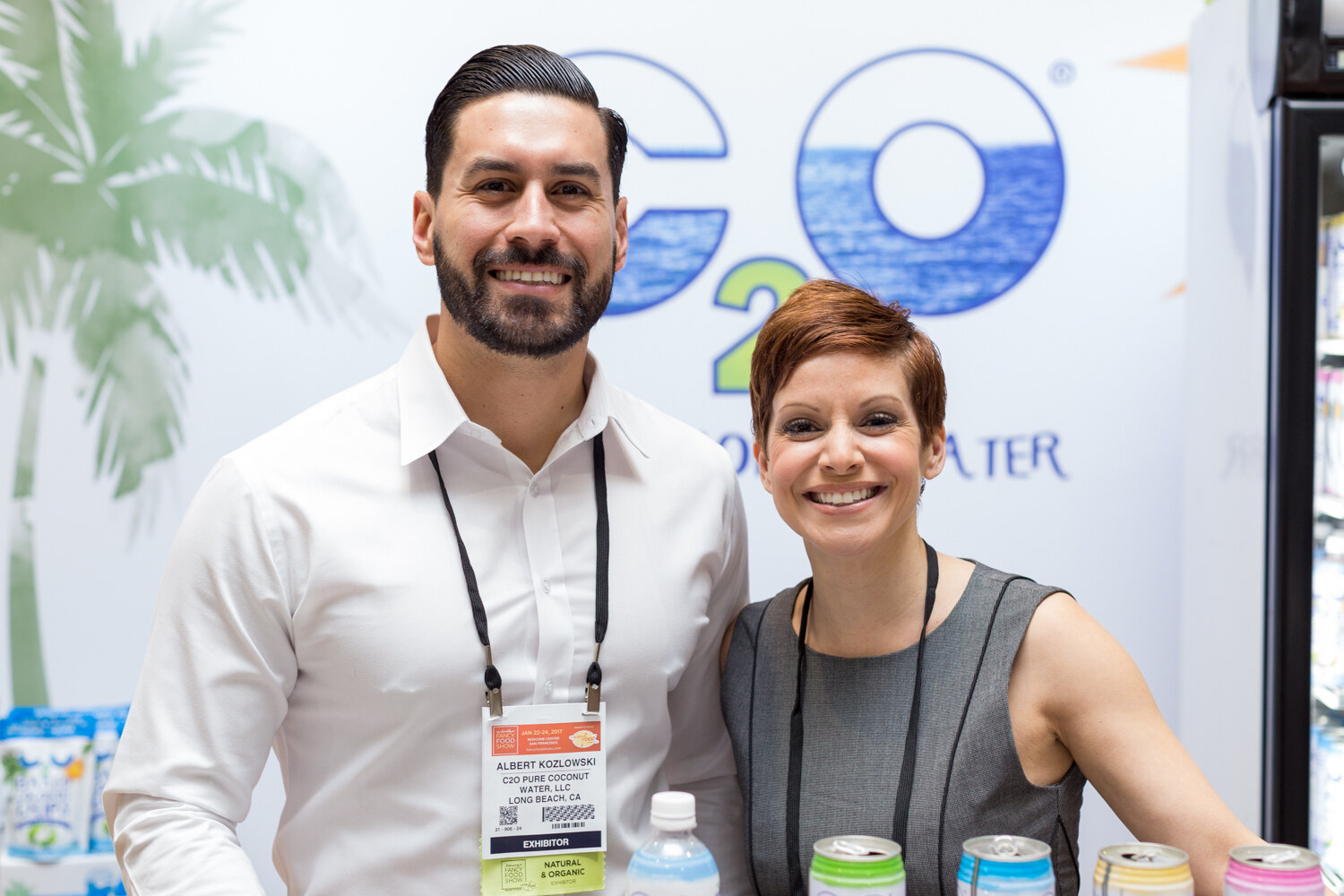 A man in a white shirt and a woman in a gray top stand in front of a C2O coconut water backdrop with cans and bottles at the bottom.
