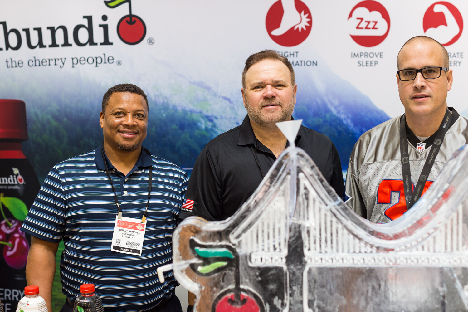 Three men stand behind an ice sculpture in front of a Cherribundi the cherry people banner and a bottle of cherry juice.