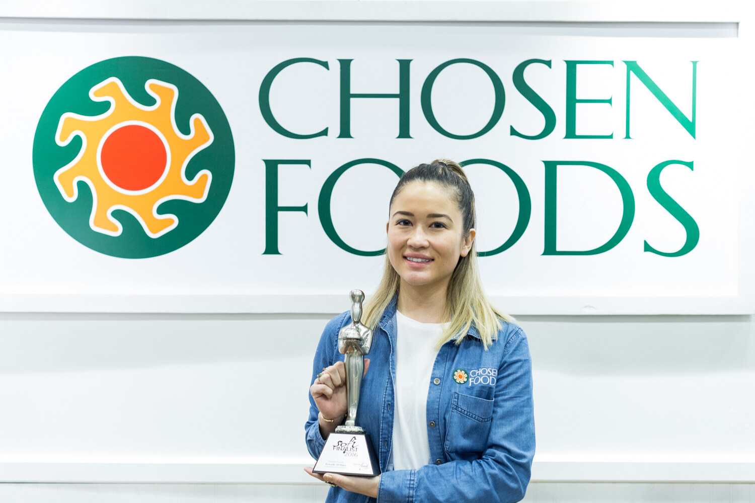 A woman in a denim Chosen Foods jacket holds a silver award trophy in front of a large Chosen Foods logo sign.