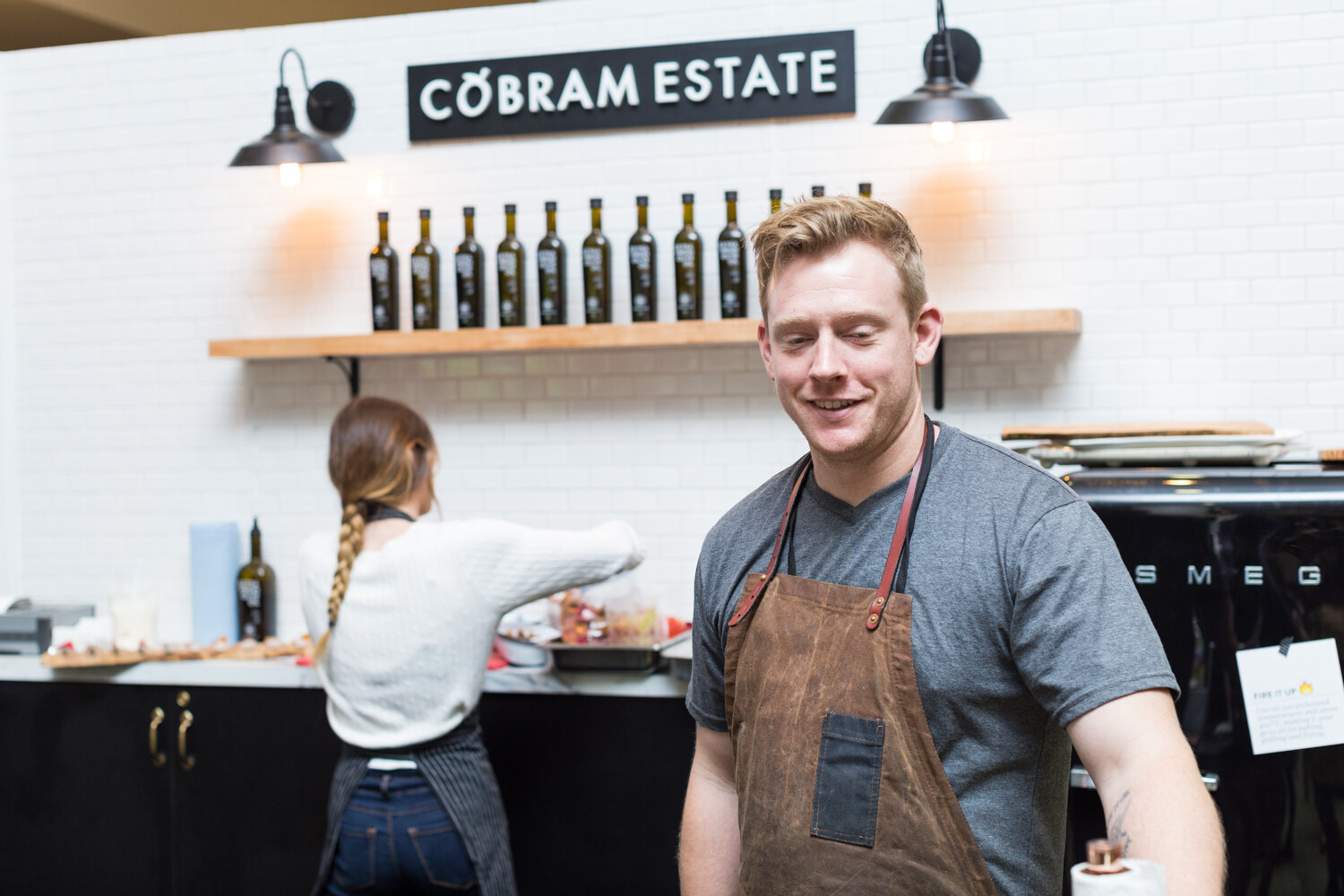 A man in a brown apron stands in front of a white tile wall with a Cobram Estate sign and a shelf of olive oil bottles while a colleague works behind him.