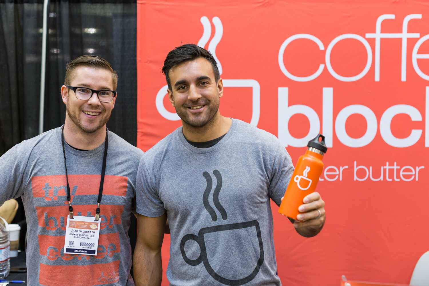 Two men in gray t-shirts stand in front of an orange Coffee Blocks banner, one holding up an orange water bottle.