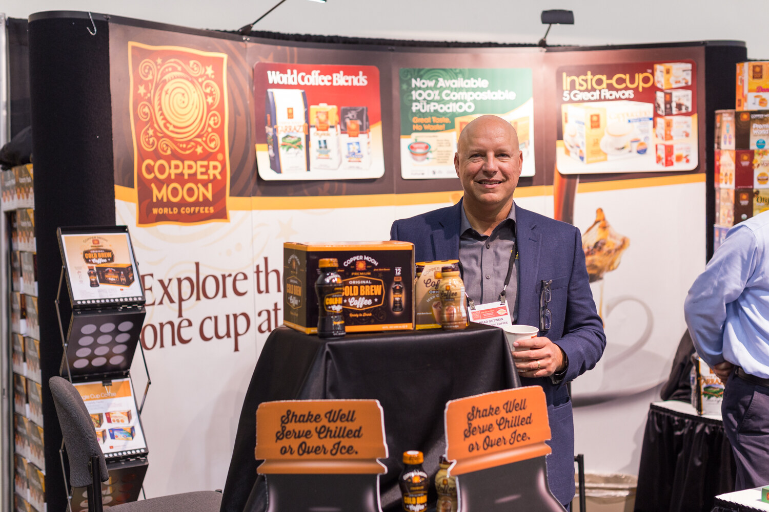 A smiling bald man in a navy blazer stands behind a table of Copper Moon cold brew bottles in front of a Copper Moon World Coffees backdrop.