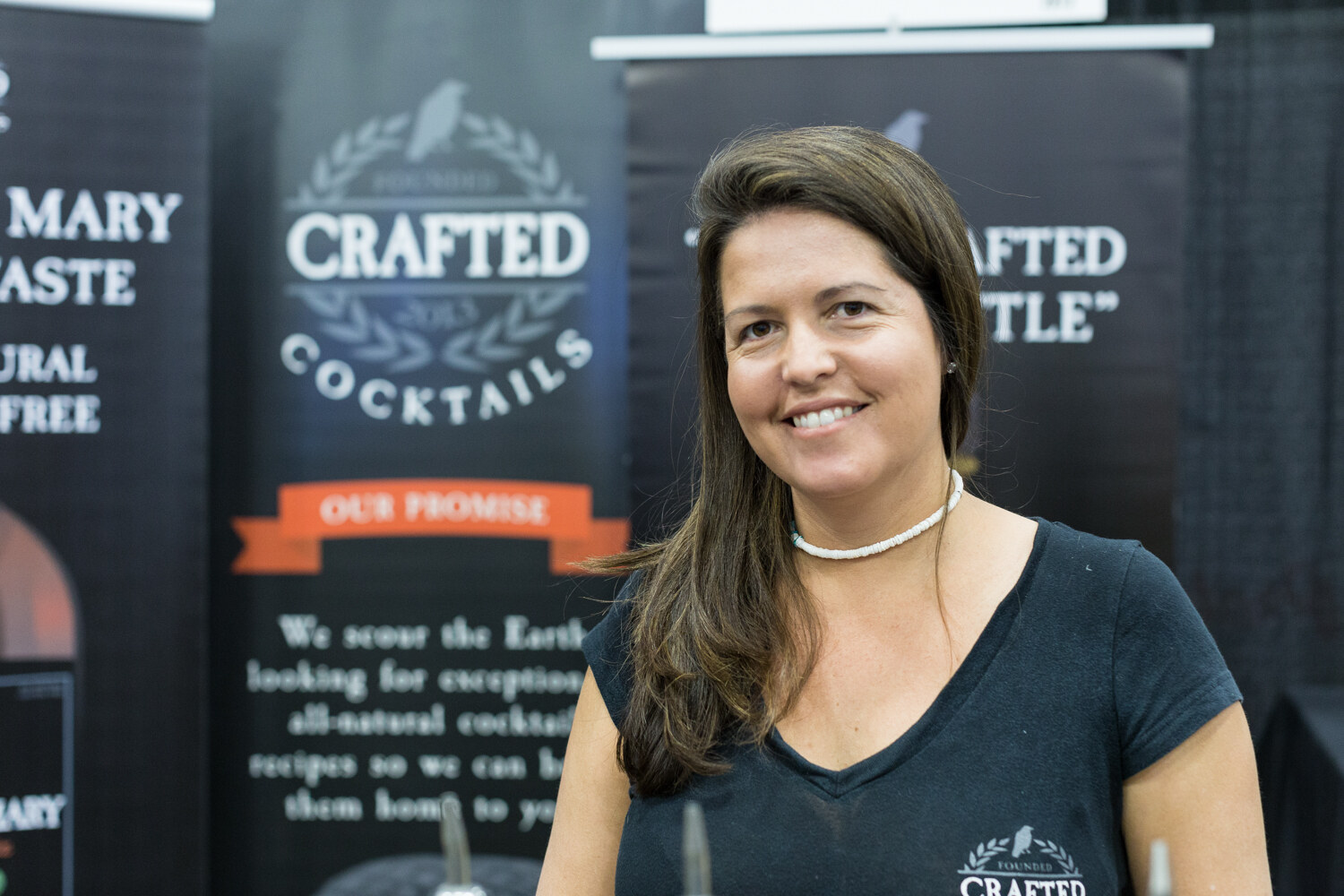 A woman in a dark shirt smiles in front of black banners reading Crafted Cocktails and Our Promise.