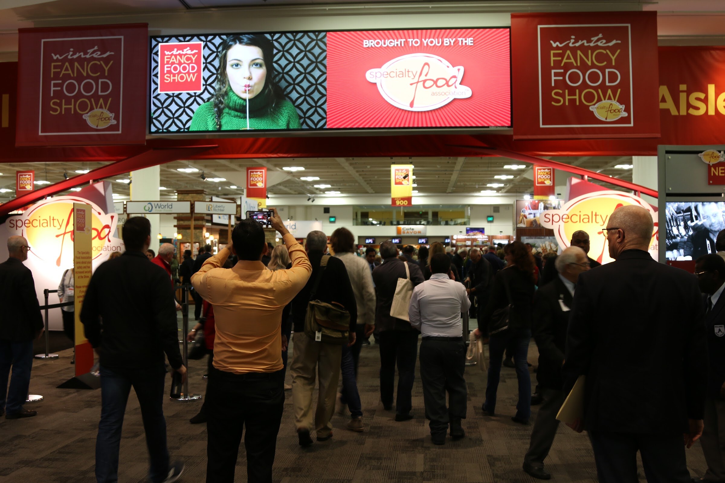 A crowd walks under red Winter Fancy Food Show banners and a video screen reading Brought to you by the Specialty Food Association, with a man in an orange shirt holding up a phone.