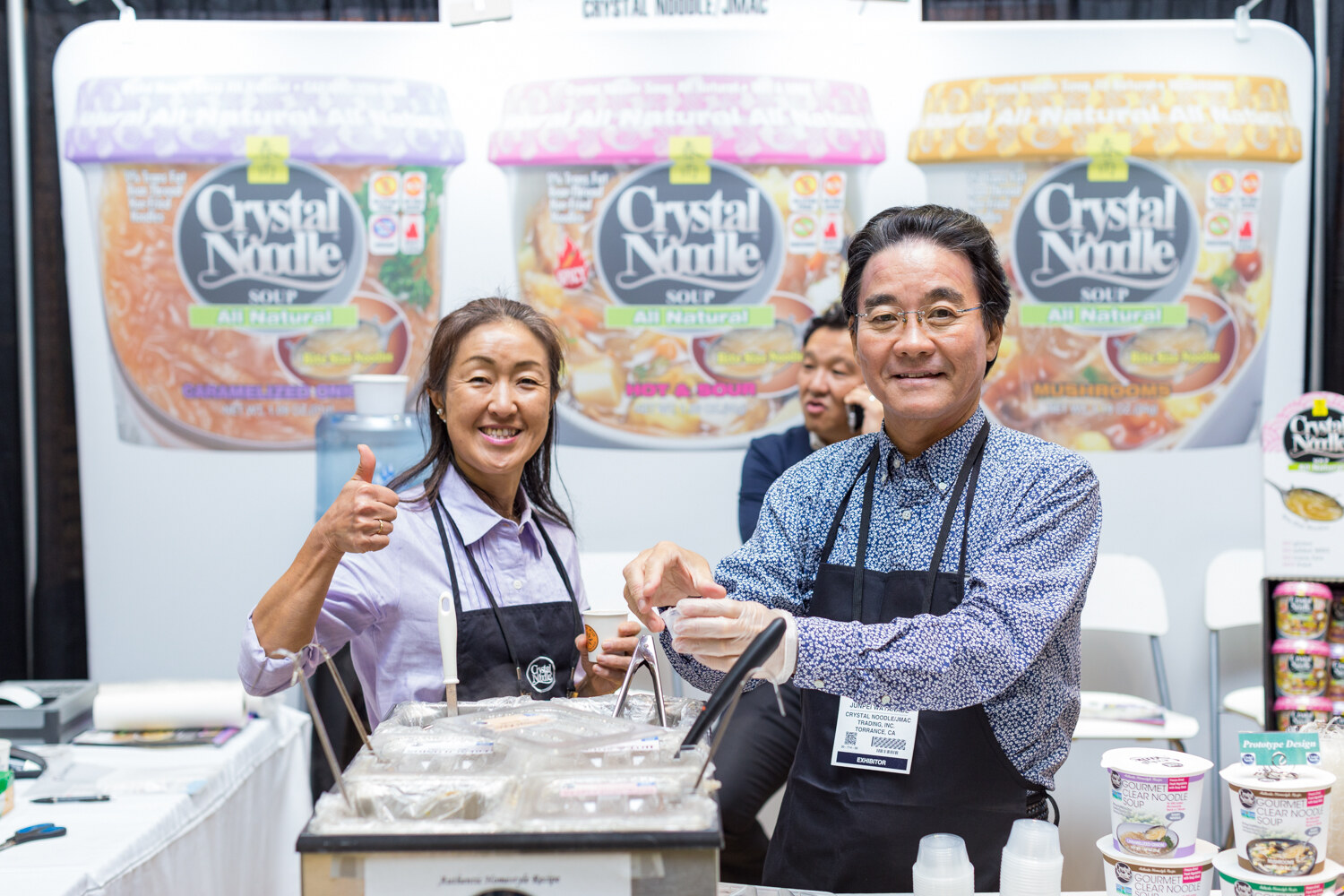 A woman giving a thumbs up and a man in an apron serve food from a steam table in front of Crystal Noodle soup graphics.