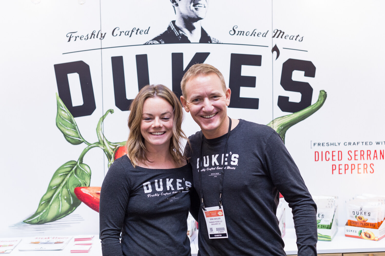 A woman and a man in Duke's long-sleeve shirts stand in front of a large Duke's backdrop with a pepper illustration.