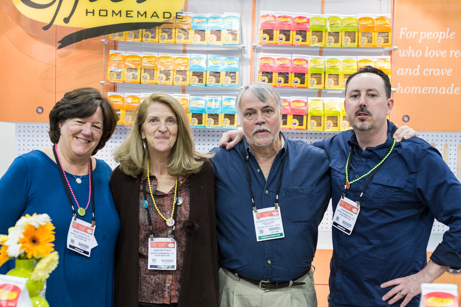 Two women and two men with exhibitor badges stand arm in arm in front of shelves of orange, yellow and blue packages under an Effie's Homemade sign.