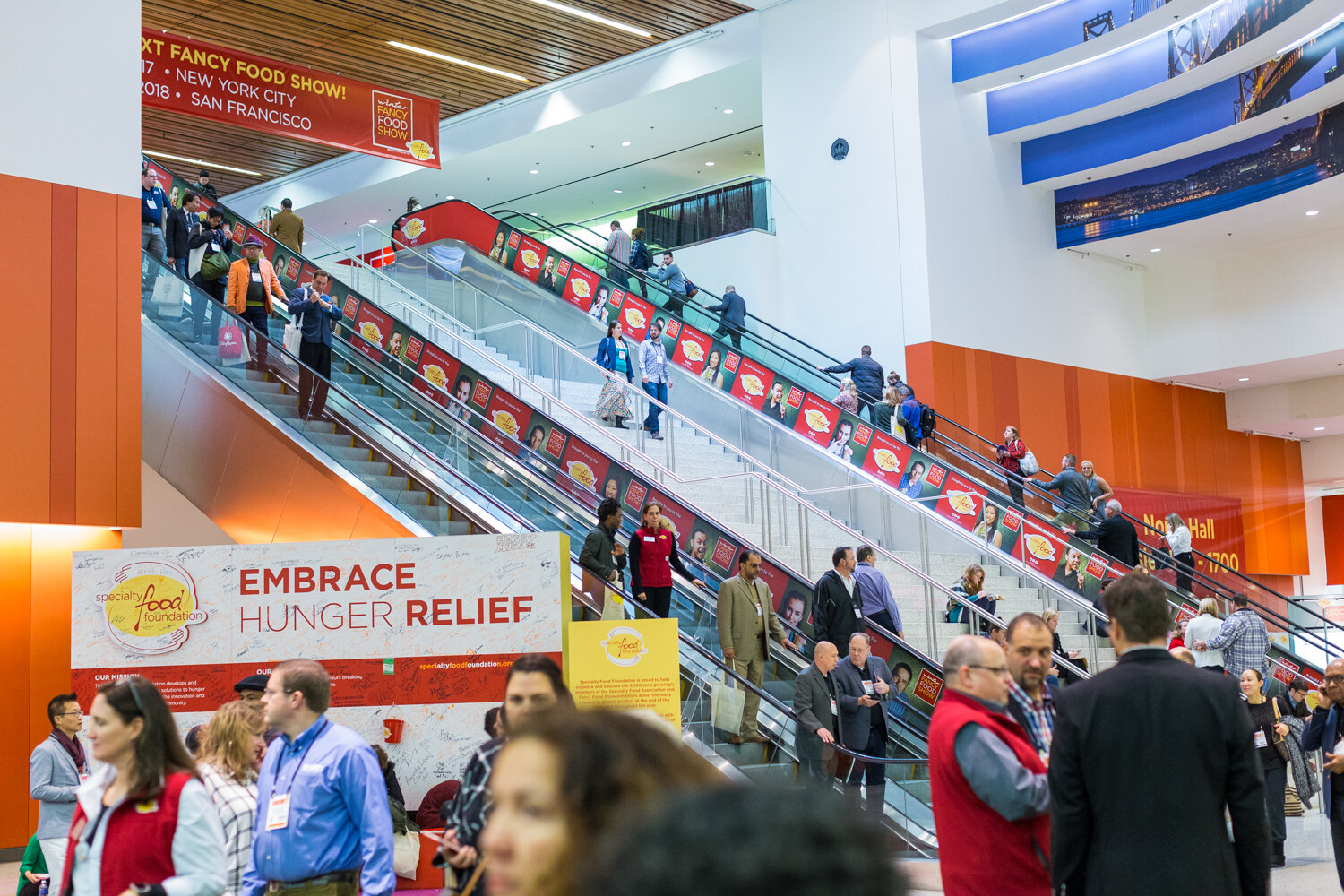 Two banks of escalators decorated with Winter Fancy Food Show banners above people in the lobby, with a sign reading Embrace Hunger Relief at lower left.