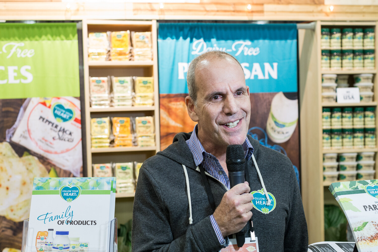 A man in a gray hoodie holds a microphone in front of shelves of dairy-free cheese slices, a Dairy-Free Parmesan banner and Follow Your Heart brochures.