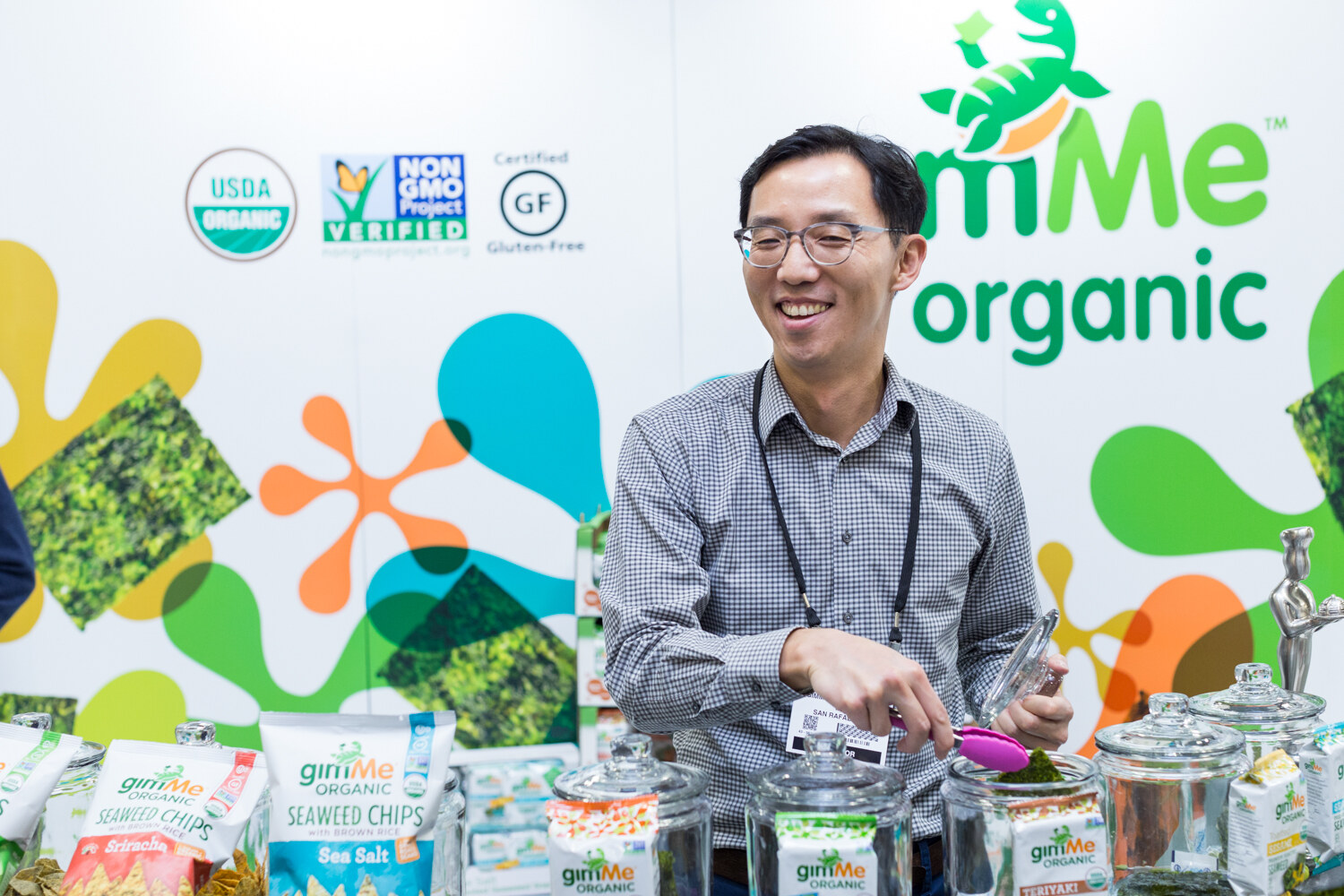 A smiling man uses tongs at a table of jars and bags of gimMe Organic seaweed chips and snacks in front of a colorful gimMe Organic backdrop.