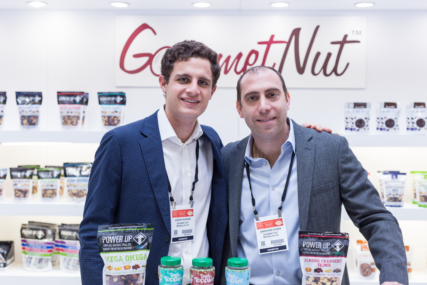 Two men with exhibitor badges stand under a Gourmet Nut sign holding bags of Power Up Mega Omega and Almond Cranberry Crunch trail mix.