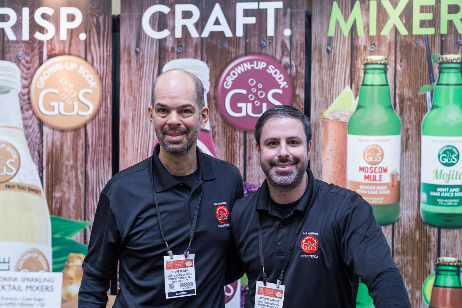 Two men in black GuS Grown-Up Soda polo shirts stand in front of a wood-look backdrop with GuS logos and bottles of Moscow Mule and Mojito soda.