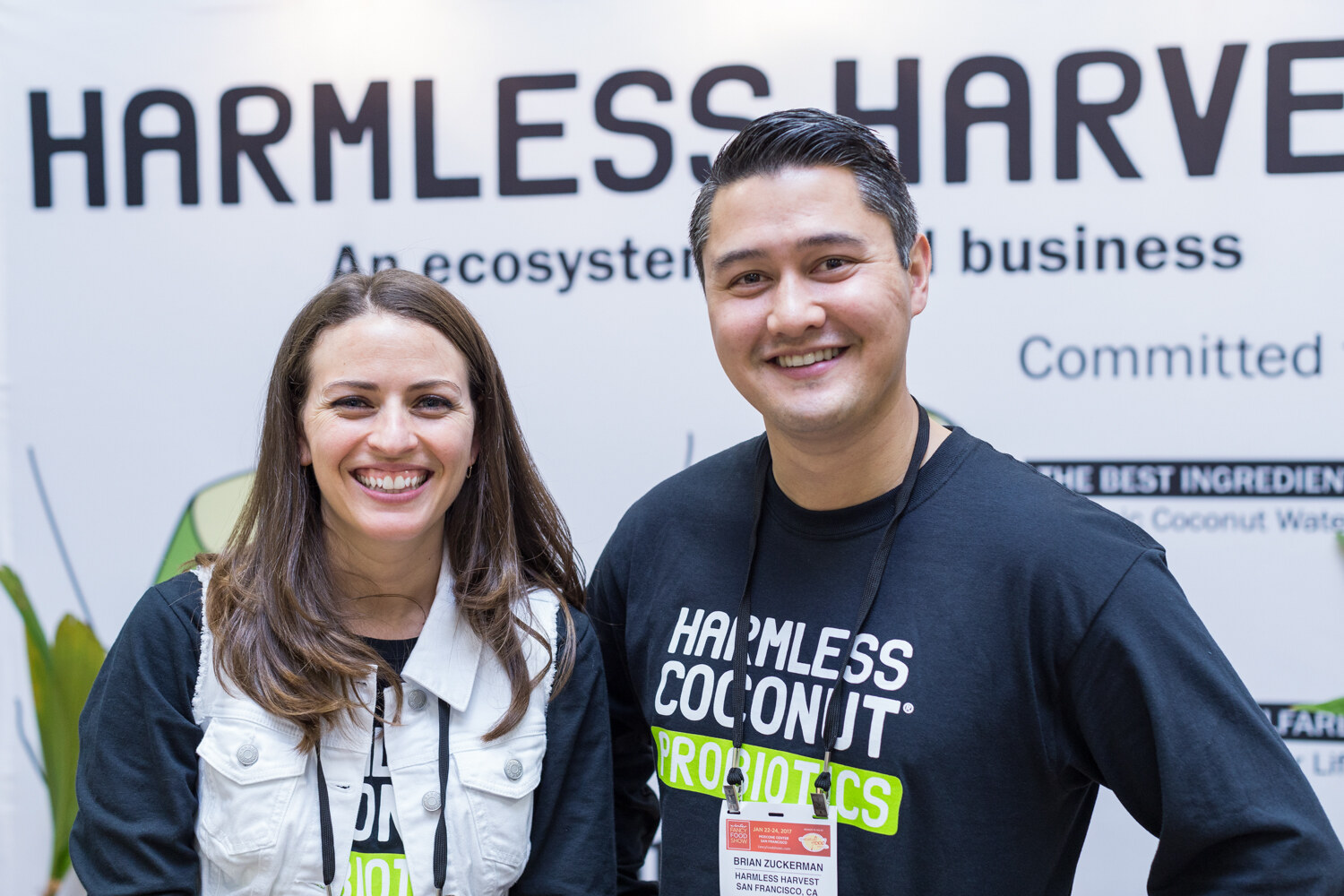 A smiling woman and a man in a Harmless Coconut Probiotics shirt stand in front of a banner reading Harmless Harvest.