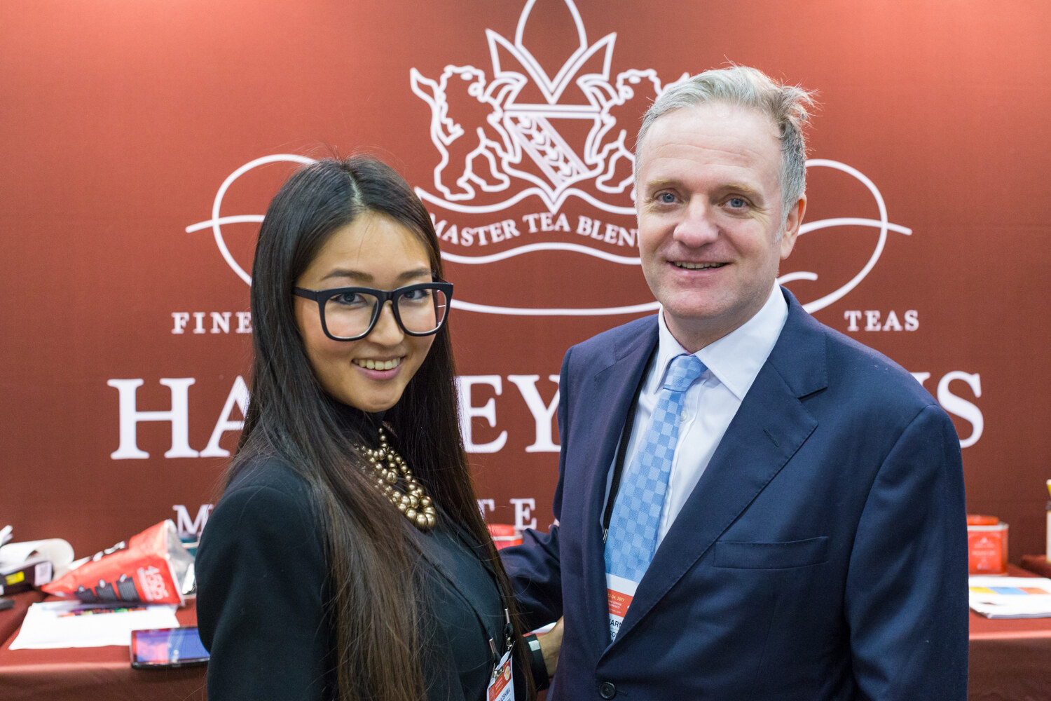 A woman in glasses and a man in a navy suit stand in front of a maroon banner with a lion crest and the name Harney.
