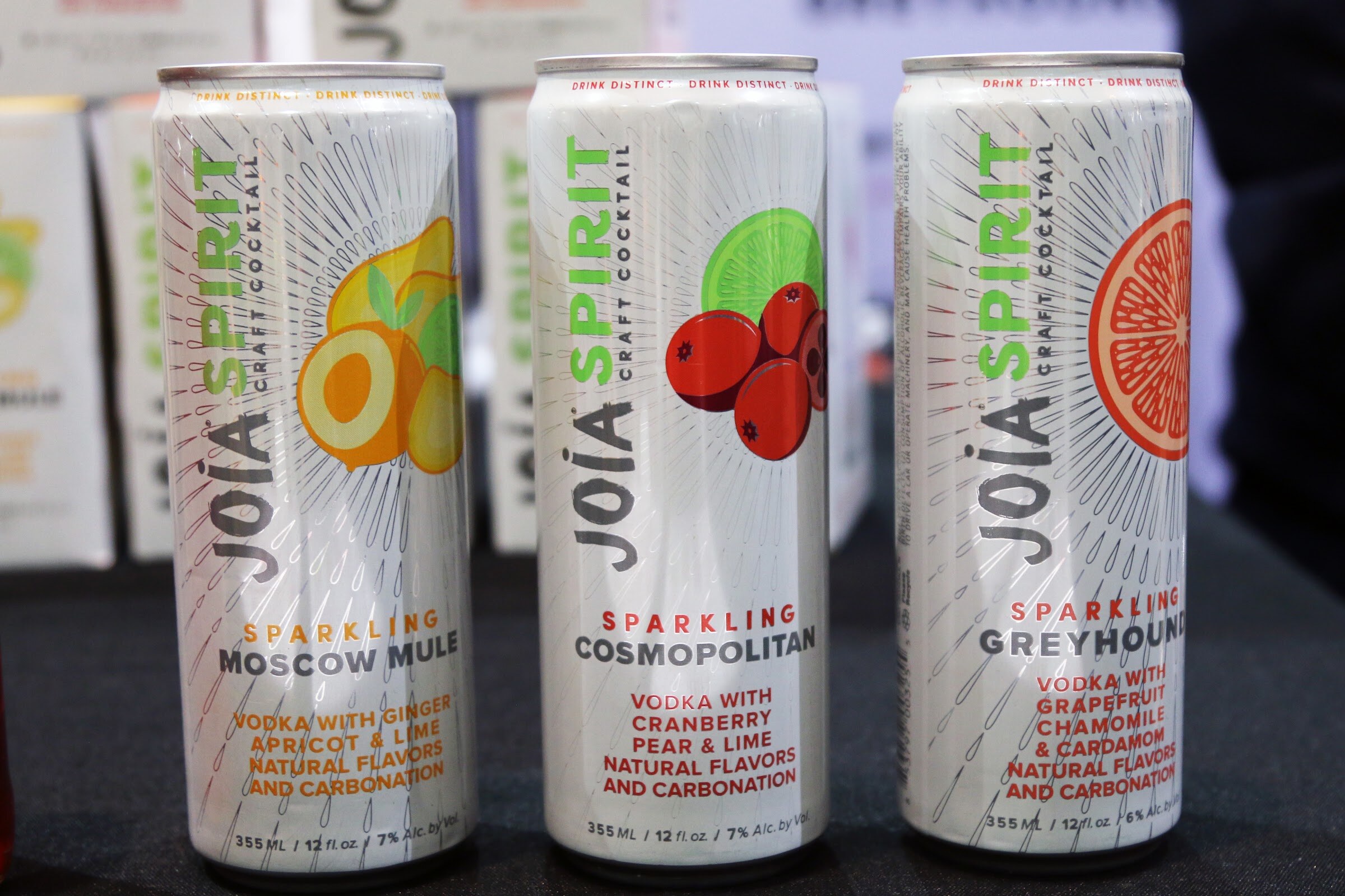 Three tall cans labeled Joia Spirit Sparkling Moscow Mule, Cosmopolitan and Greyhound on a dark table.