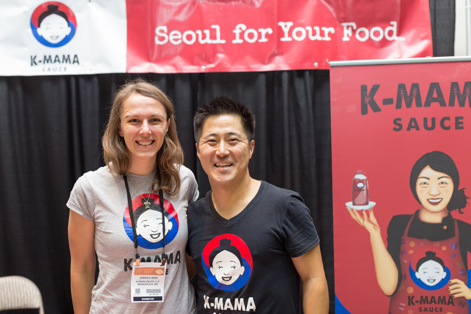 A woman and a man in K-Mama shirts smile in front of a red K-Mama Sauce banner reading Seoul for Your Food.