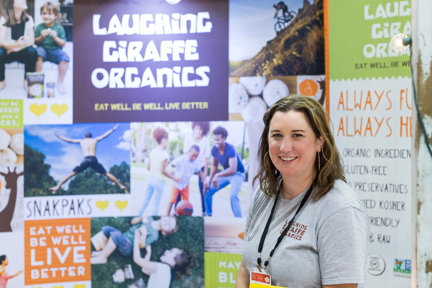 A woman in a gray Laughing Giraffe Organics t-shirt stands in front of a collage of lifestyle photos and a Laughing Giraffe Organics sign.